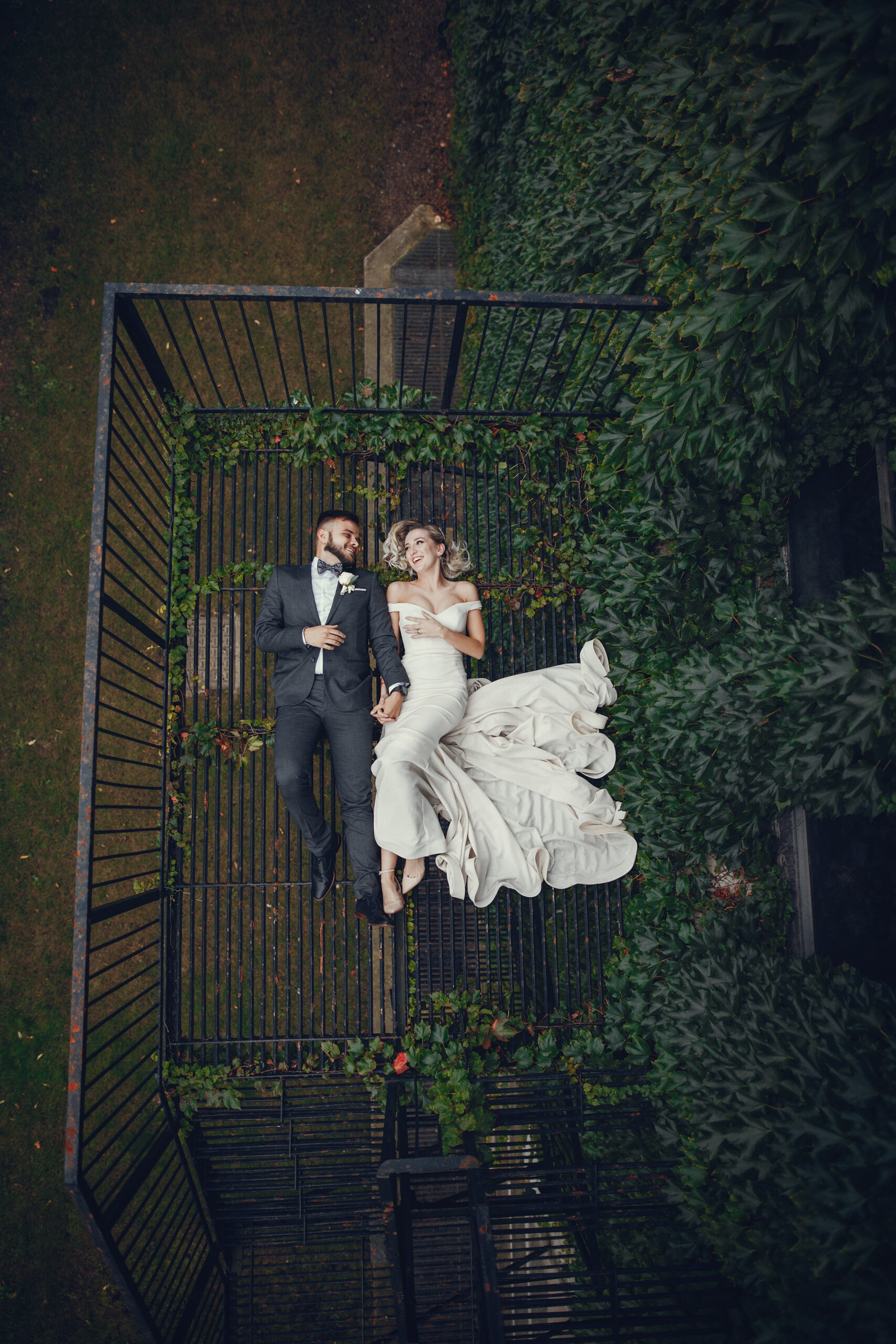 Beautiful bride in a long white dress. Handsome groom in a black suit. Couple lie on a metal
