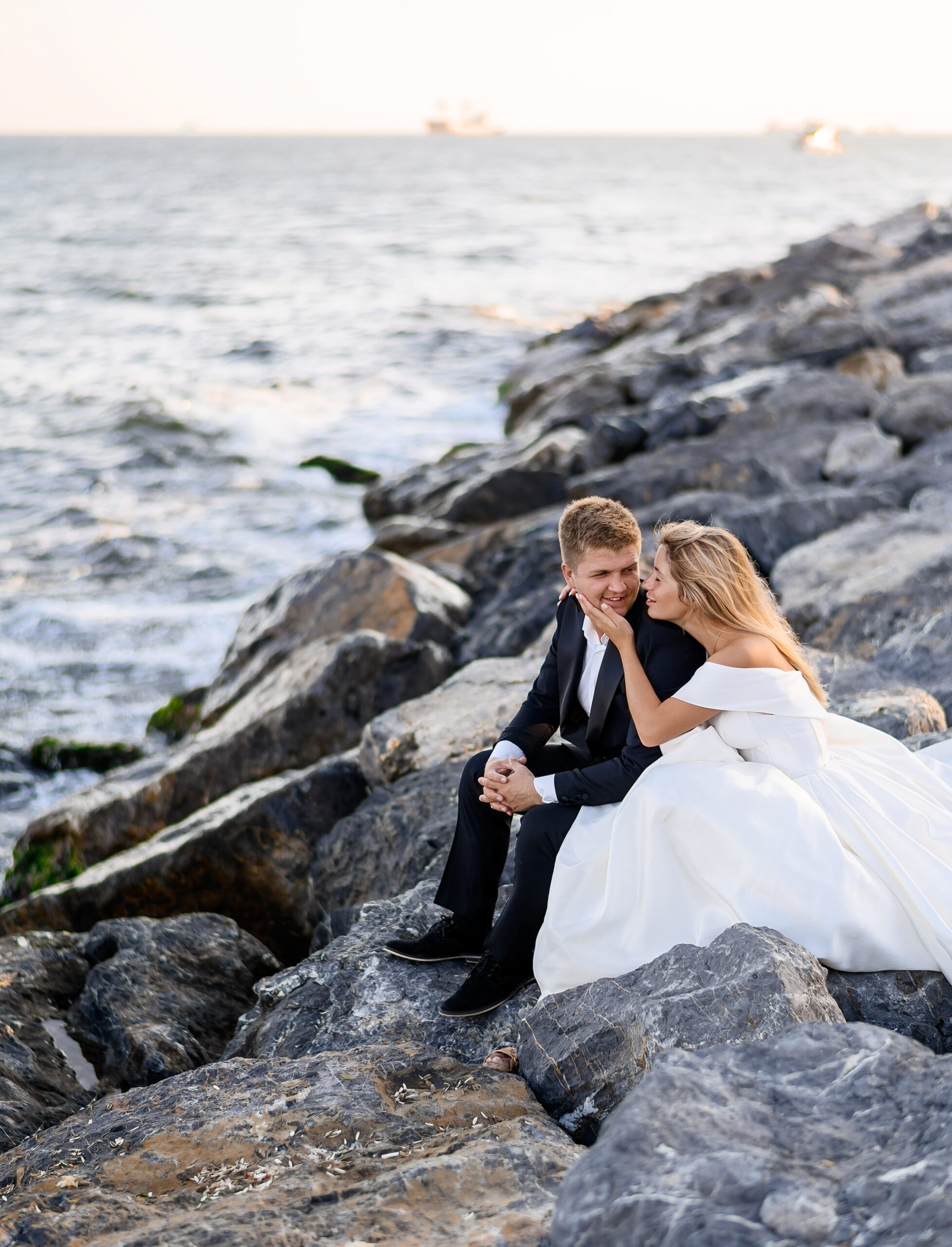 Side view of couple sitting on stone beach near sea, bride in wedding dress touching face of groom in stylish black suit looking at each other and enjoying nature landscapes